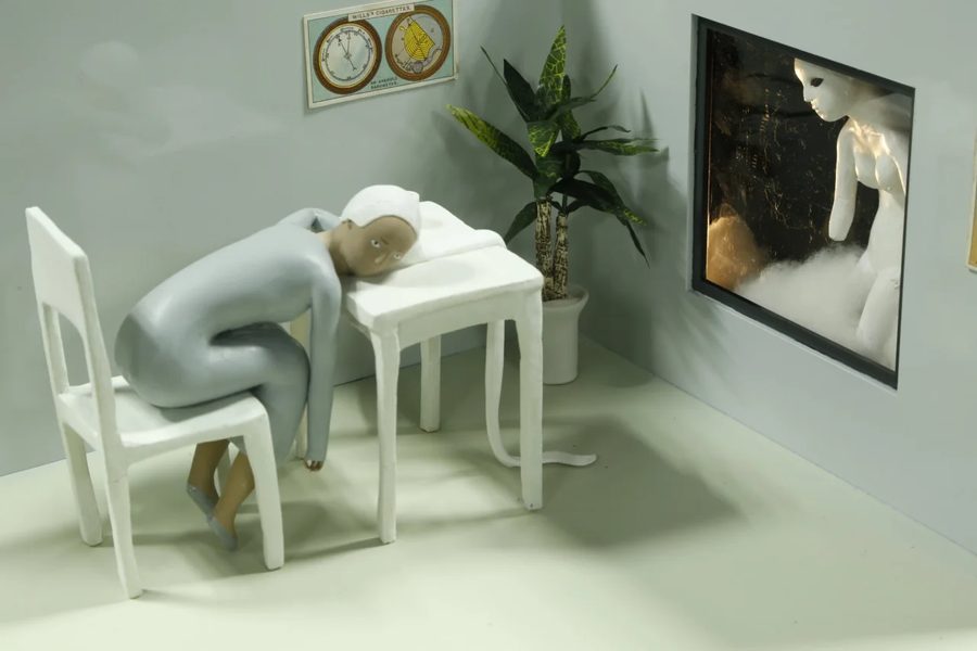 A pale figure slumped over a white table next to a potted pl, 2021